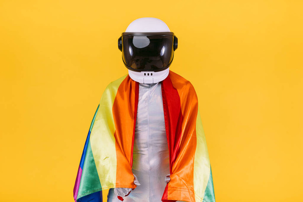 Kasklı ve gümüş takım elbiseli bir astronot gibi giyinmiş eşcinsel adam sarı arka planda lgtbi topluluğunun bayrağını taşıyor. Gay, homoseksüel, trans, hak ve cinsiyet gururu kavramı. - Fotoğraf, Görsel