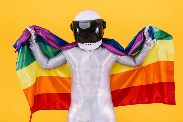 ヘルメットと銀のスーツを着た宇宙飛行士として身を包んだゲイの男性は、黄色の背景にlgtbi集団の旗を持っています。ゲイ、同性愛、トランス、権利、ジェンダーの誇りの概念. - 写真・画像