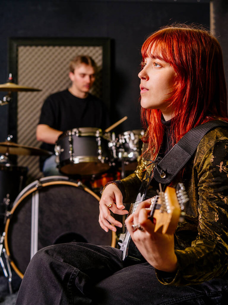 Young redhead woman playing electric guitar and a male drummer in a recording studio. - Photo, image