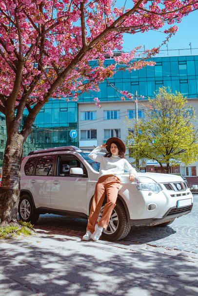 κομψό χαμογελώντας όμορφη γυναίκα στέκεται κοντά στο αυτοκίνητο ανθίζοντας δέντρο sakura στο φόντο άνοιξη έρχεται - Φωτογραφία, εικόνα