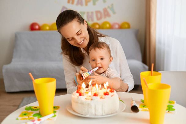 Portret van moeder met grappige baby vieren eerste verjaardag, taart is verrassing voor kind, doe een eerste wens, blazen partij hoorn, poseren thuis met ballonnen en partij inscriptie op de achtergrond. - Foto, afbeelding
