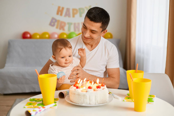Portrait of father with adorable toddler baby celebrating first birthday, cake is surprise for child, make a first wish, posing at home with balloons and party inscription on background. - Foto, afbeelding