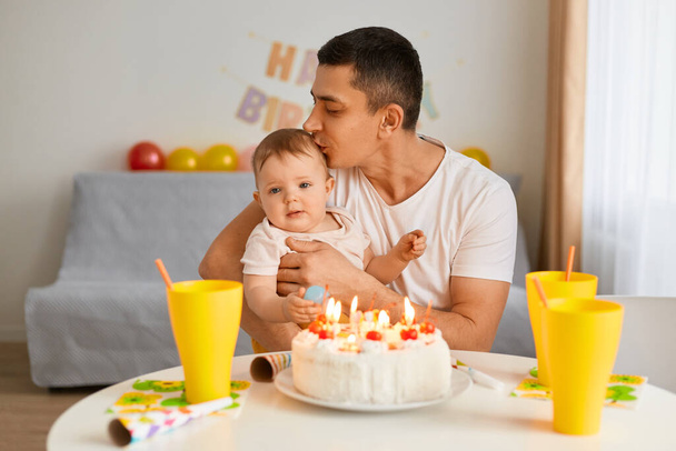 Indoor shot of brunette female man with infant baby celebrating first birthday, sitting at table with birthday cake and drink, father kissing his infant daughter, celebrating together. - Photo, image