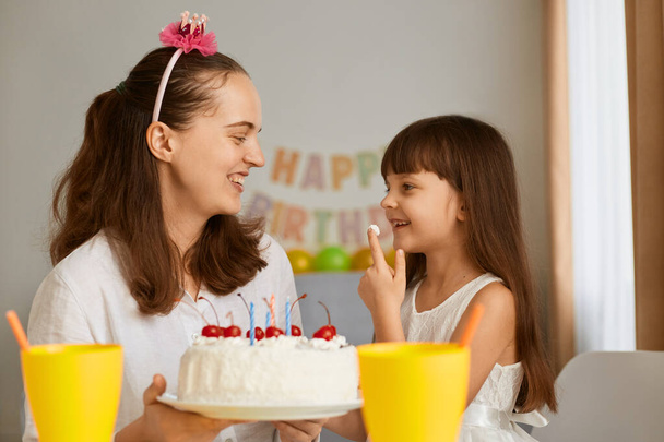 Portrait of charming little girl receiving congratulations from lovely mother at home, loving mom giving cake with candles to excited happy daughter during Birthday celebration. - Фото, изображение