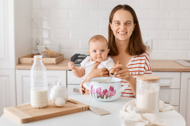 Portrait of smiling dark haired woman sitting at table surrounded with different ingredients for baking, satisfied female expressing happiness, spending time together with her child. - 写真・画像