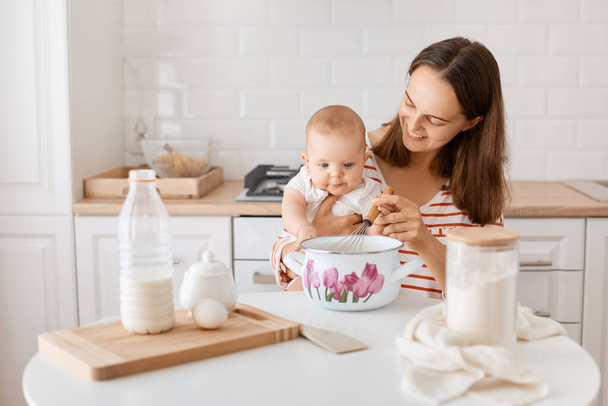 Portrait of happy delighted woman with dark hair sitting at table with infant baby daughter, mommy holding whisk together with kid, cooking bakingin kitchen. - Fotó, kép