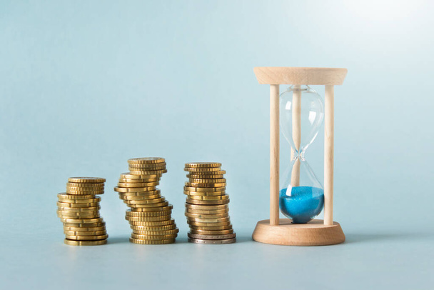 Money accumulation, investment concept - three stacks of gold coins with small sand clock on blue background. - Foto, immagini
