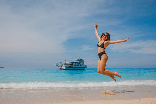 heureuse femme souriante en maillot de bain noir sautant à la mer croisière sur la plage petit navire sur fond - Photo, image