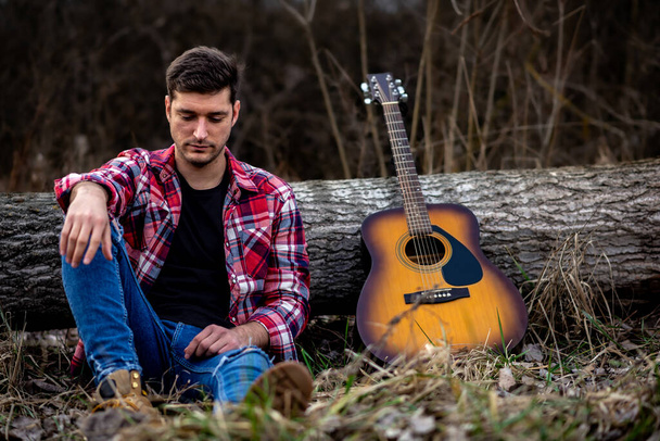 A handsome young man is sitting on ground leaning on a fallen tree next to an acoustic guitar in a nature park. - Foto, Imagem