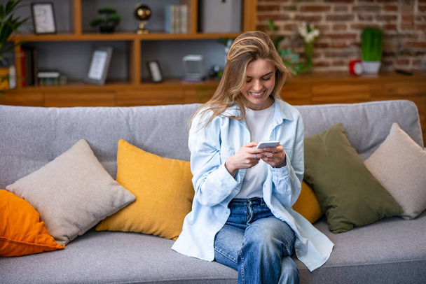 Smiling young woman using mobile phone app playing game, shopping online, ordering delivery relax on sofa. - Foto, immagini
