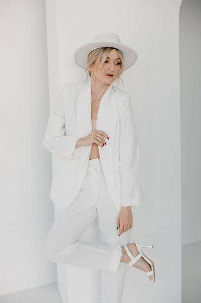 Confident young blond woman smiling, looking at camera isolated on white background. Studio portrait of successful friendly female in white suit and hat, posing over white wall. - Foto, imagen