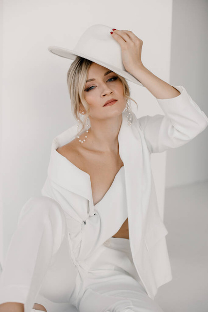 Confident young blond woman smiling, looking at camera isolated on white background. Studio portrait of successful friendly female in white suit and hat, posing over white wall. - Photo, image