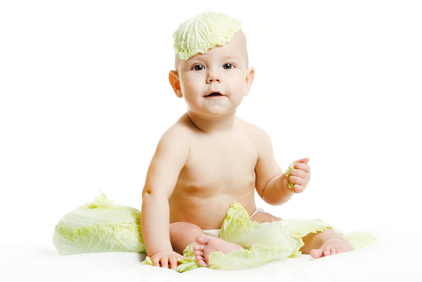 Naked baby in diaper is playing with fresh organic vegetables and fruits isolated on a white background. Малыш кусает свежую зеленую голову капусты. Здоровое питание для детей. - Фото, изображение