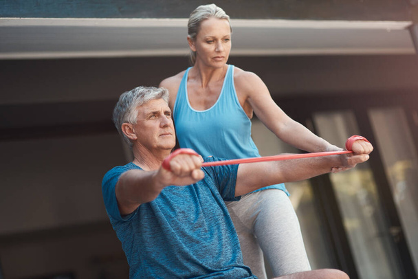 Center yourself and you can do anything. Shot of a mature man stretching his arm muscles with an elastic band while being assisted by a woman. - Photo, Image