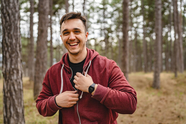 Egy férfi boldog fiatal kaukázusi férfi mosolyog, miközben arra készül, hogy az erdőben vagy a nyilvános parkban edz, kapucnis pulcsit vesz fel vagy vesz le, igazi emberek utánozzák a helyet. - Fotó, kép