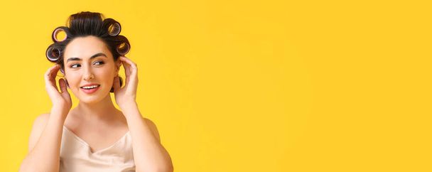 Beautiful young woman in pajamas and hair curlers on yellow background with space for text - Fotografie, Obrázek