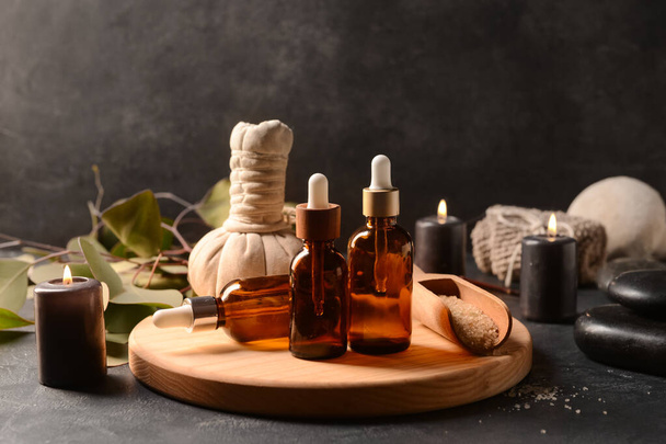 Tablero de madera con botellas de aceite esencial, sal marina, bolsa de masaje y velas ardientes sobre fondo oscuro - Foto, imagen