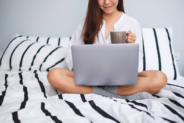 自宅の白い居心地の良いベッドに座っている間にコーヒーを飲みながら、ラップトップコンピュータで使用して作業する美しい女性のクローズアップイメージ - 写真・画像