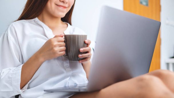 Nahaufnahme einer schönen Frau, die mit Laptop arbeitet und Kaffee trinkt, während sie zu Hause auf einem weißen, gemütlichen Bett sitzt - Foto, Bild