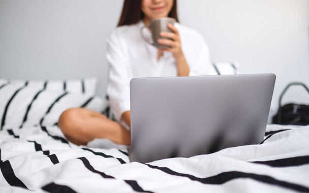Immagine in primo piano di una bella donna che utilizza e lavora sul computer portatile, bevendo caffè mentre si siede su un letto bianco accogliente a casa - Foto, immagini