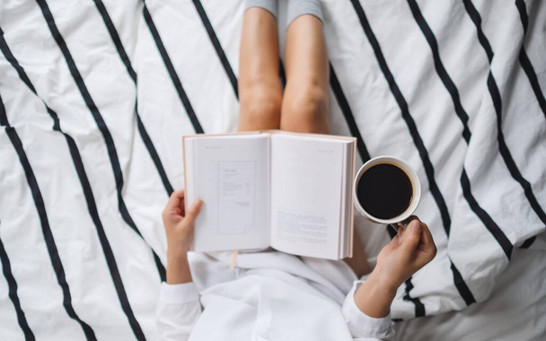 Top view image of a woman reading book and drinking hot coffee in a white cozy bed at home in the morning - Photo, Image