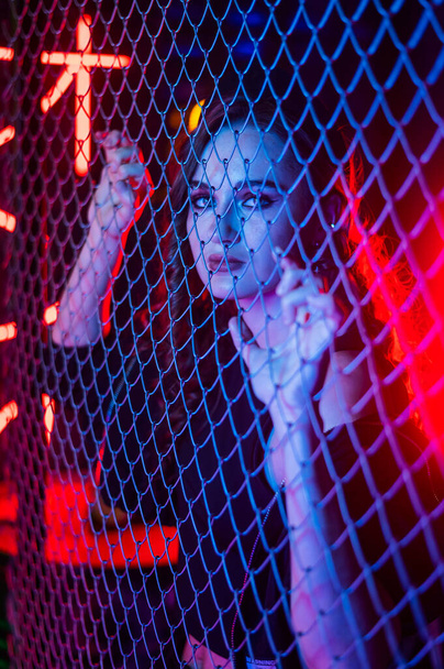 Caucasian woman in neon studio behind chain-link mesh.  - Photo, Image