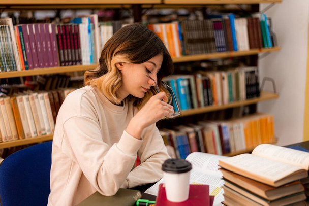 Piękna dziewczyna studiuje na biurku w bibliotece zakładając okulary - Zdjęcie, obraz