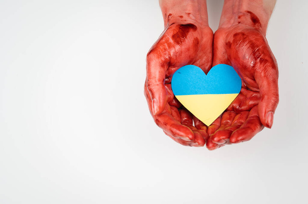 Woman with hands covered in blood holding a heart with the flag of ukraine on a white background.  - Foto, Imagem