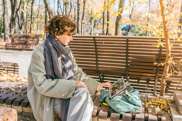 teenager girl with curly brown hair, sitting on bench in autumn city park, chats online with friends or family. Positive emotion on face, communication, video conference, internet friend - Photo, Image