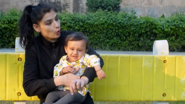 Egy gyönyörű bébiszitter, aki egy édes csecsemőt tart a karjában - családi kötelék, rugalmasság, kemény élet. Egy gondoskodó indiai anya megnyugtatja boldogtalan kislányát, miközben ül egy padon a parkban - egy fiatal szülő, sír baba - Felvétel, videó