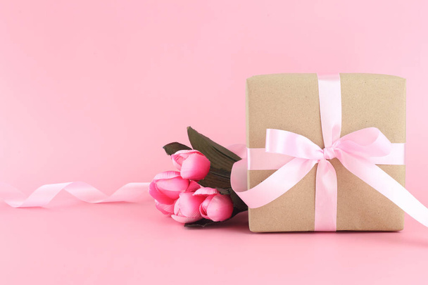 Καφέ χάρτινο κουτί δώρου με ροζ σατέν σγουρά κορδέλα φιόγκο σε παστέλ ροζ φόντο με όμορφα λουλούδια. Ημέρα της μητέρας, Ημέρα του πατέρα, Ημέρα του Αγίου Βαλεντίνου, έννοιες γενεθλίων με χώρο αντίγραφο. - Φωτογραφία, εικόνα