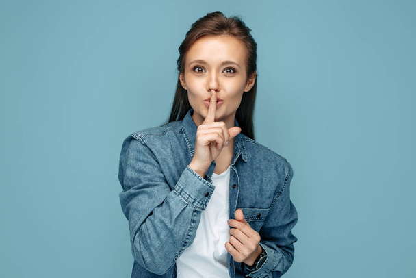 Young woman in jeans shirt showing keep silent gesture looking to camera over blue background. - Photo, image