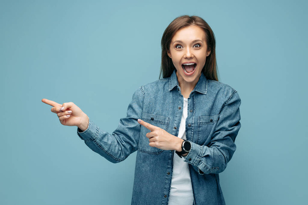 Excited young woman in jeans shirt pointing fingers away over blue background. Look in camera. - Zdjęcie, obraz