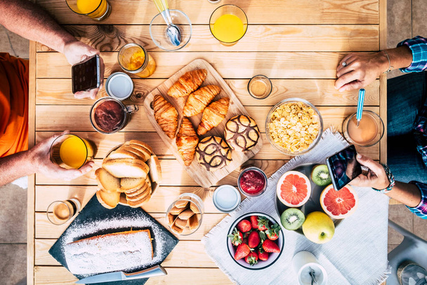 Vertical view of table full of breakfast food and group of people eating and enjoying it using mobile phone. Family and friends eat together and enjoy morning leisure time. Bakery and fruit - Foto, Imagem