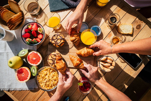 Vertical view of table full of breakfast food and group of people eating and enjoying it using mobile phone. Family and friends eat together and enjoy morning leisure time. Bakery and fruit - Zdjęcie, obraz