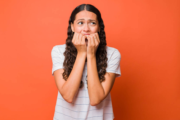 Scared and anxious young woman biting her nails while feeling afraid against an orange background - Photo, image
