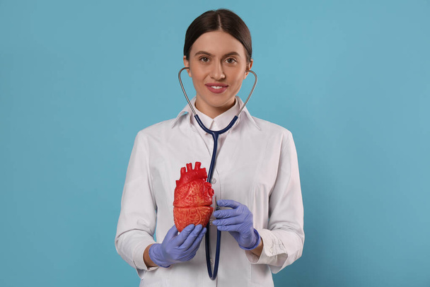 Doctor with stethoscope and model of heart on light blue background. Cardiology concept - Фото, зображення