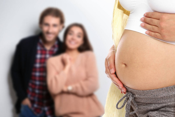 Surrogacy concept. Young pregnant woman and blurred view of happy couple on light background - Фото, зображення