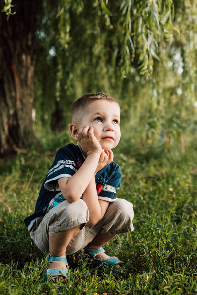 A little boy crouched under a tree, his hand propping up his head and angry - Photo, Image