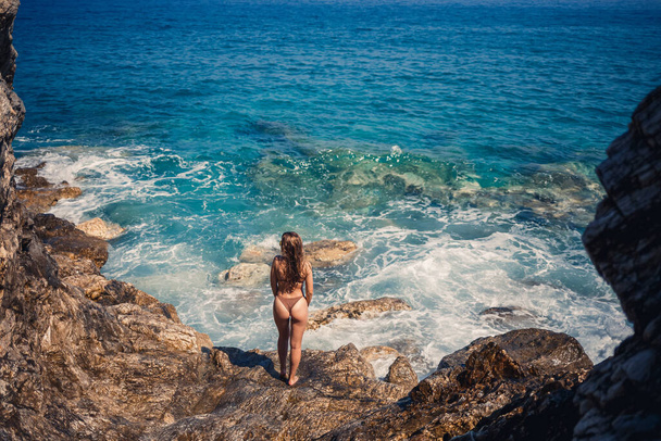 Donna sexy in costume da bagno completo con lunghi capelli cammina su grandi rocce su una spiaggia rocciosa durante una tempesta in mare. Vista posteriore. Donna costumi da bagno isola tropici paesaggio esotico passeggiata - Foto, immagini
