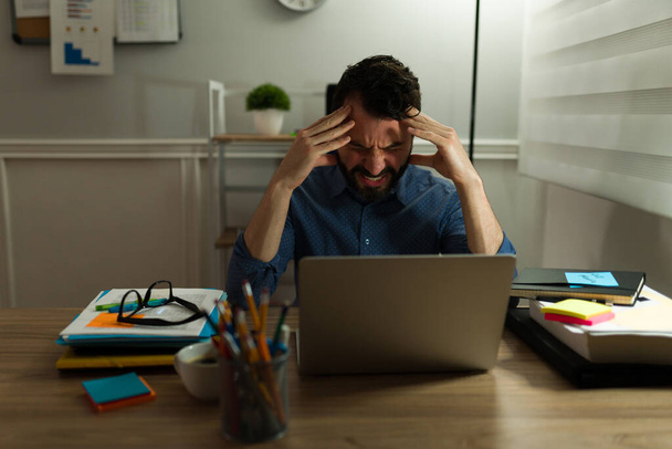 Ελκυστικός άνθρωπος που υποφέρει από έντονο πονοκέφαλο λόγω της δουλειάς του και αισθάνεται εξαντλημένος στο γραφείο - Φωτογραφία, εικόνα