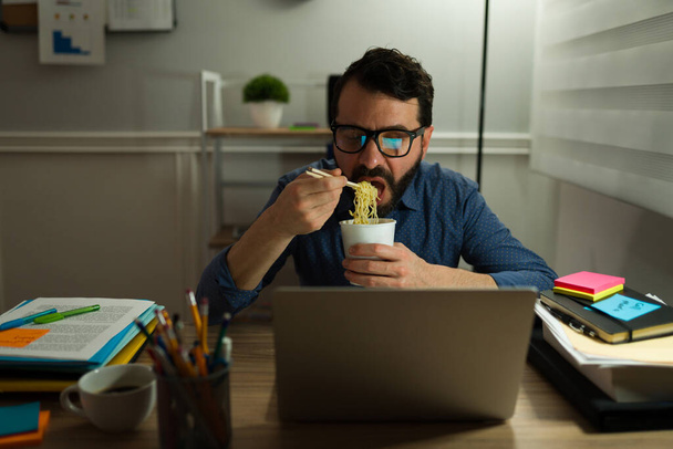 Uomo caucasico maniaco del lavoro che mangia zuppa di tagliatelle davanti al computer mentre lavora fino a tardi in ufficio - Foto, immagini