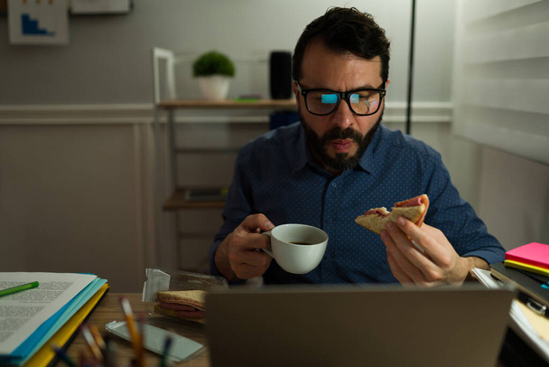 Workoholik muž s brýlemi pomocí notebooku při jídle sendvič a pití kávy při práci pozdě  - Fotografie, Obrázek