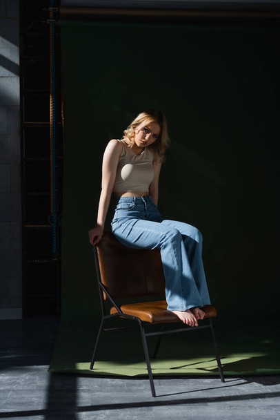 full length view of young barefoot woman in jeans and top posing on chair on dark background - Photo, Image