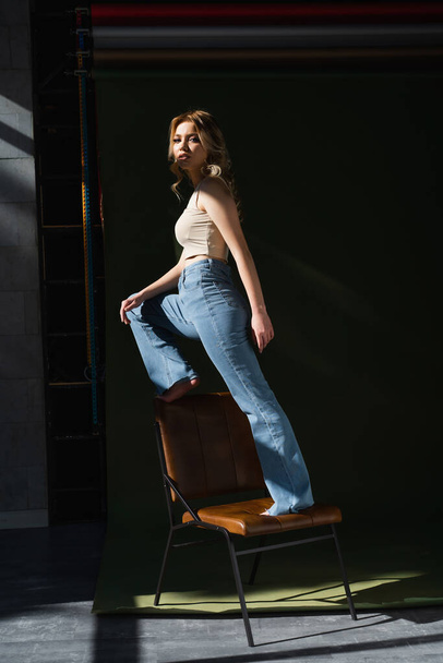 volle Länge der barfüßigen Frau in Jeans, die in die Kamera schaut, während sie auf einem Stuhl vor dunklem Hintergrund steht - Foto, Bild