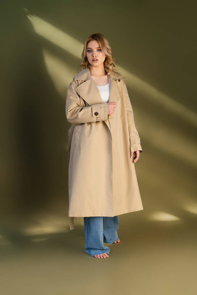 full length of young barefoot woman in oversize trench coat on beige background with lighting - Photo, Image