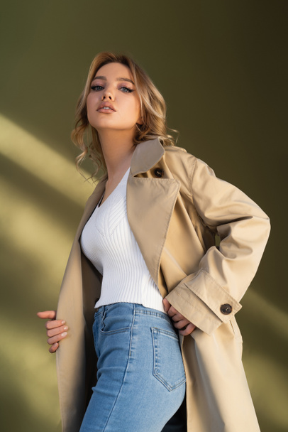 low angle view of pretty woman in trench coat and jeans looking at camera on beige background - Fotó, kép