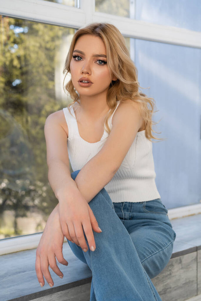 young woman in jeans sitting near window and looking at camera - Foto, Imagem