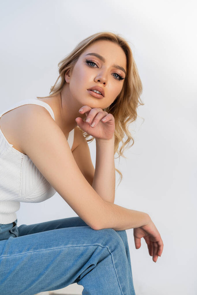 pretty woman in jeans looking at camera while sitting with hand near face isolated on white - Zdjęcie, obraz