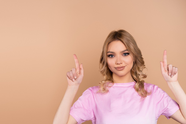 smiling woman in t-shirt pointing up with fingers isolated on beige - Foto, Imagen